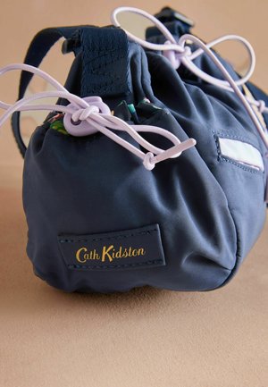 Marineblauer Stoffbeutel mit Kordelzugverschluss, versehen mit fliederfarbenen Schnüren und einem gold bestickten Markenetikett. Glatte Textur mit gerafftem Design.