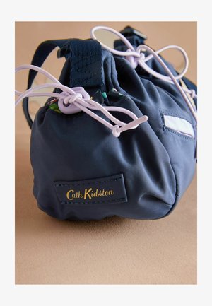 Marineblauer Stoffbeutel mit Kordelzugverschluss, versehen mit fliederfarbenen Schnüren und einem gold bestickten Markenetikett. Glatte Textur mit gerafftem Design.