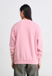 Ροζ φούτερ με στρογγυλή λαιμόκοψη, ραμμένα μανίκια και στρίφωμα. Διαθέτει μαλακή υφή και χαλαρή εφαρμογή. Συνδυάζεται με μπλε τζιν παντελόνι.