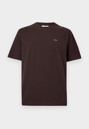 Jeune homme debout portant un t-shirt marron foncé, un pantalon large beige et des chaussures noires sur un fond clair uni.