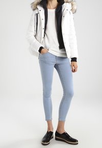 Bílá padded bunda s černým lemováním a umělou kožešinou na kapuci, v kombinaci s lehkým svetr, modrými skinny džíny a černými lakovanými slip-on botami.