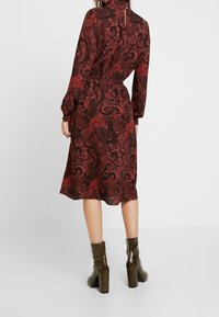 Vestido en estampado paisley rojo y negro con mangas largas, cintura elástica y cuello alto. Combinado con botas de tobillo brillantes con estampado de cocodrilo.