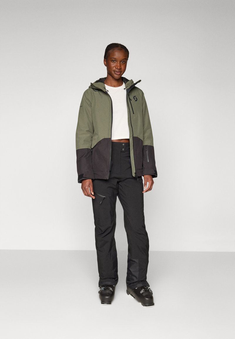 Grüne und schwarze Skijacke mit Kapuze, Reißverschlusstaschen und Logodetail, kombiniert mit schwarzen Schneehosen und Stiefeln. Glatte Stofftextur.