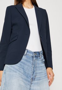 Marinblå blazer med en knapp, två framfickor och en skräddarsydd passform, kombinerat med en vit ribbad topp och en ljus tvättad denimkjol.