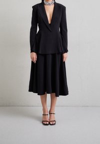 Černý oblekový kabát s hlubokým véčkovým výstřihem, kombinovaný s volnou černou sukní. Disponuje elegantními liniemi a strukturovaným siluetou. Černé sandály na tenkých podpatcích.