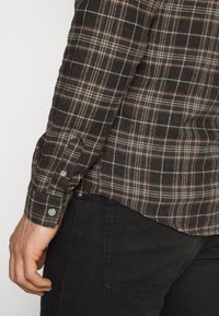 Hihna ruskeassa ruudullisessa paidassa, jossa on napitettavat hihansuut, lepää käden vieressä, yhdistettynä mustiin farkkuihin, joissa näkyy takatasku.