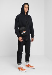 Felpa con cappuccio nera, jeans scuri, sneakers arancioni con suola bianca e una borsa a tracolla nera con un logo bianco e rosso.