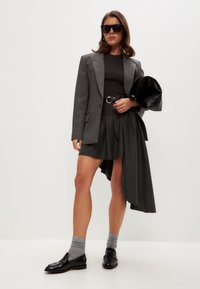 Grå skräddarsydd blazer över en svart topp, kombinerad med en högmidjad asymmetrisk grå kjol. Svarta loafers och grå strumpor fulländar looken.