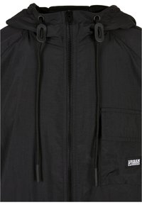 Veste noire avec capuche, fermeture éclair à l'avant, cordons ajustables et une poche poitrine avec un tag logo. Tissu léger avec une finition texturée.