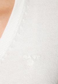 Close-up van een witte gebreide trui met geribbelde V-hals en subtiel reliëf GANT-logo op de borst.
