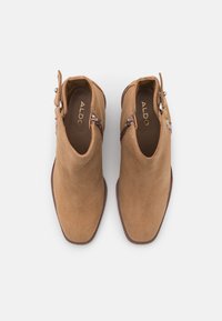 Botas de tobillo de ante color tan con punta afilada. Presenta una cremallera lateral y una hebilla decorativa. Textura suave con acentos mínimos.