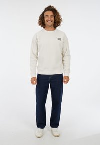 Crème sweatshirt met ronde hals en ribgebreide manchetten, voorzien van een klein logopatch. Gecombineerd met donkerblauwe rechte spijkerbroek en witte sneakers.
