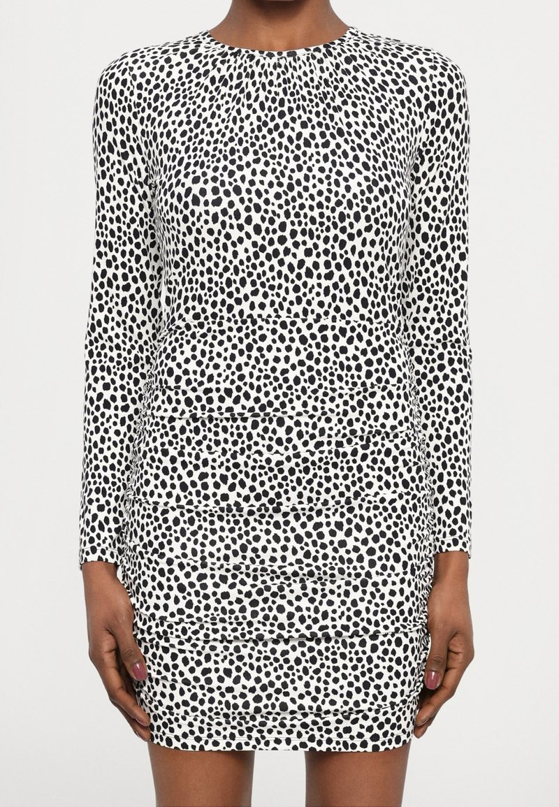 Femme portant une robe ajustée à manches longues, noire et blanche à pois, avec un col rond et un détail de jupe à volants.