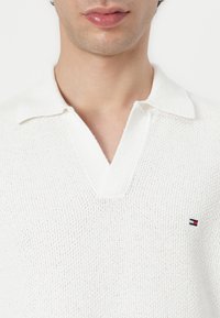 Vit stickad polo tröja med V-hals, med ett strukturerat mönster och en liten flerfärgad logotyp på vänster bröst.