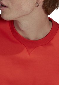 Ung person med ljus hy och fräknar som bär en röd t-shirt med rund halsringning och en liten hoops örhänge, vänd åt vänster.