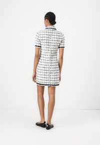White and black tweed mini dress with short sleeves, collar, and frayed hem. Paired with black flat shoes. Rear view, minimal background.