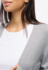 Hellgrauer Cardigan über einem weißen Oberteil, mit weicher Strickstruktur und offenem Vorderteil, sichtbaren Ärmeln und dezenten Nähdetails.