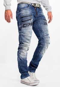 Homme portant un jean bleu délavé avec plusieurs poches zippées et des baskets blanches, se tenant devant un fond uni.