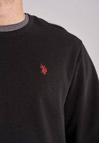 Sweatshirt noir en coton, avec un col ras du cou. Logo rouge brodé notable au niveau de la poitrine gauche. Texture lisse.