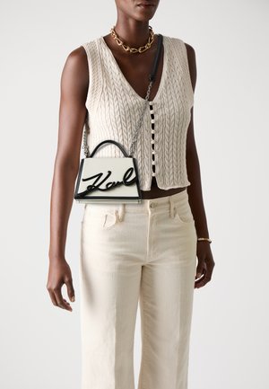 Sac à main blanc avec une signature noire, design structuré, poigné supérieur et bandoulière en chaîne détachable. Le modèle porte un gilet en tricot beige à cable et un pantalon clair.