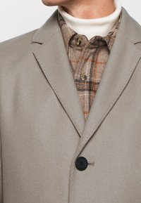 Cappotto di lana beige con colletto a notched, chiusura con bottoni e dettagli in bottone neri, indossato sopra una camicia di flanella a quadri marrone.