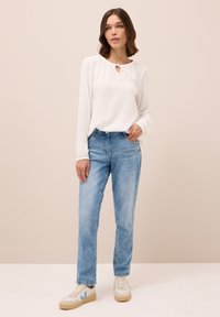 Blusa bianca a maniche lunghe con dettaglio a cravatta sul collo; abbinata a jeans in denim azzurro chiaro. Il modello indossa sneakers color crema e azzurro chiaro.