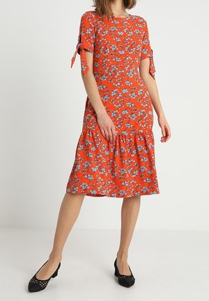 Abito rosso floreale con maniche corte, caratterizzato da dettagli con fiocco alle maniche, orlo arricciato e una vestibilità ampia. Abbinato a scarpe nere.