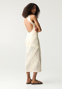 Knie-langes cremefarbenes Kleid mit einem strukturierten Spitzenmuster, einem offenen Rücken und diagonalen Streifen aus kontrastierendem Stoff. Getragen mit braunen Sandalen.