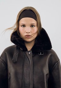 Giacca di pelle marrone con un alto e morbido colletto in pelliccia nera. Presenta una tasca con zip, grandi bottoni e un design strutturato. Il modello ha i capelli castani.