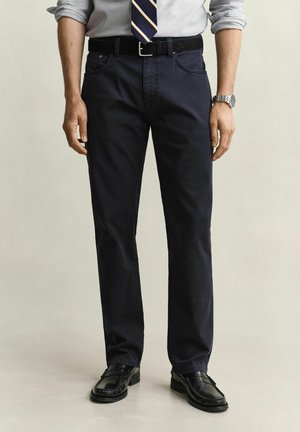 Man draagt donkerblauwe broek, zwarte riem, zwarte leren schoenen, grijs overhemd met opgerolde mouwen, gestreepte stropdas en zilveren polshorloge, staand op een lichte achtergrond.