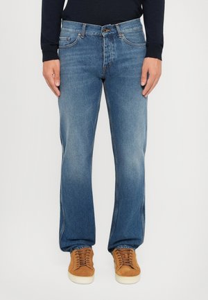 Front view of person wearing medium blue straight-leg jeans and tan suede sneakers with white soles against white background.