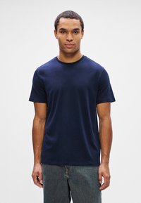 Homem de frente, vestindo uma t-shirt simples de mangas curtas azul-escura e jeans cinzas, contra um fundo cinza-claro.