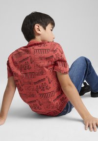 Raudona marškiniai su grafikų atvaizdais, juodais tekstais ir iliustracijomis, derinami su mėlynomis džinsais ir juodais batais, sėdima pozicija.