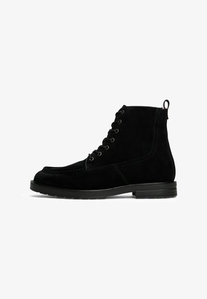 Stivale alla caviglia in suede nero con lacci, fascetta per il tacco e tacco basso impilato, vista laterale su sfondo bianco.