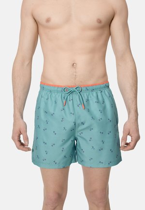Hombre sin camiseta que lleva pantalones de baño turquesa con pequeños estampados de libélulas azules marinas y una cintura con cordón naranja, de pie frente a un fondo liso.