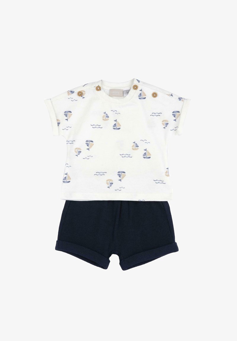 Maglietta bianca per bambino con stampe di barche a vela blu e beige e bottoni in legno sulle spalle, abbinata a pantaloncini blu scuro semplici.