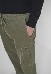 Pantalones de pana verde con una superficie texturizada, que cuentan con una cintura ajustable con cordón y un bolsillo lateral tipo cargo con un acento de logo.