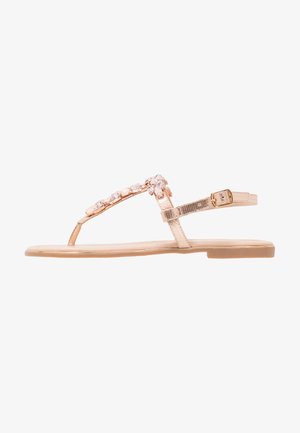 Ružičaste zlatne ravne sandale s T-trakom ukrašenim prozirnim rhinestones, sintetičkom gornjom stranom i niskim, fleksibilnim đonom. Podesivi remen oko gležnja.