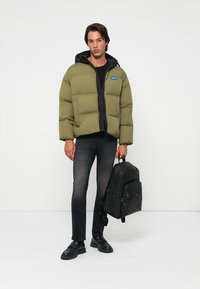 Olive green puffer jacket with a hood, black lining, and blue logo patch; paired with dark jeans and a black leather backpack.