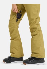 Pantaloni da neve gialli con aperture in rete con zip, caratterizzati da un taglio affusolato. Indossati con stivali neri e suole bianche in stile sneaker.