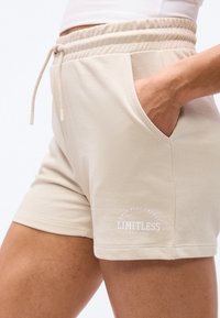 Μπεζ σορτς με ελαστική μέση και κορδόνι, πλαϊνή τσέπη και τυπωμένο κείμενο "LIMITLESS" στον αριστερό μηρό, φορεμένα με το χέρι μέσα στην τσέπη.