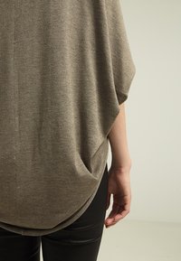 Luźny, taupe sweter z wysokim przodem i niższym tyłem, rękawami typu nietoperz oraz teksturowanym wykończeniem, zestawiony z czarnymi legginsami.