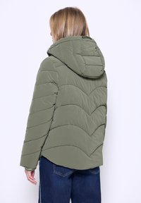 Vadderad grön jacka med huva, med ett chevron-quiltat mönster och en avslappnad passform. Materialet verkar slätt med en matt yta.