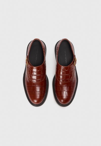 Zapatos marrones de piel sin cordones con textura de cocodrilo, puntera redondeada y un acento de hebilla dorada en el lateral. Suela de goma.