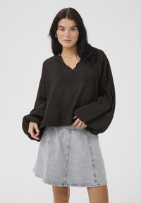 Schwarze, kurz geschnittene Bluse mit V-Ausschnitt, langen Puffärmeln und strukturiertem Finish, kombiniert mit einem hellgrauen, denim A-Linien-Rock mit Nahtdetails.