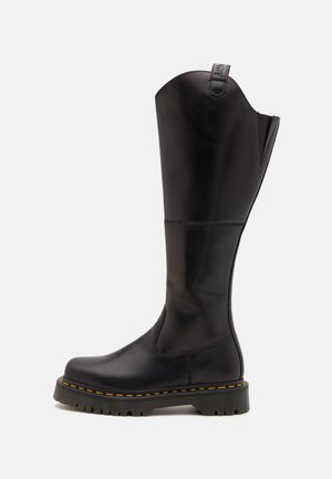 Botte noire en cuir montante jusqu'au genou avec semelle crantée épaisse et coutures jaunes le long du bord de la semelle sur fond blanc.