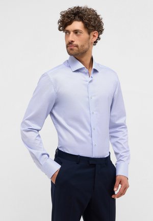 Chemise bleu clair à boutons avec un petit motif géométrique, manches longues et col classique. Portée avec un pantalon bleu marine.