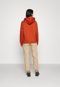 Sudadera de color óxido con capucha ajustable, puños acanalados y dobladillo. Combinada con pantalones beige claros y ajustados en los tobillos y zapatos negros.