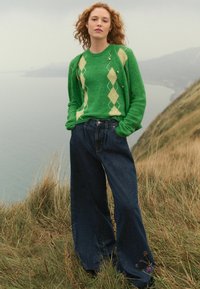 Groene, gestructureerde wollen cardigan over een groene ruitjestrui. Donkerblauwe wijde denim jeans, met een grasachtergrond en uitzichten op de oceaan.