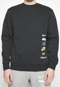 En person som bär en svart sweatshirt och grå sweatpants, båda med flera Nike-logotyper i olika stilar på högra sidan.